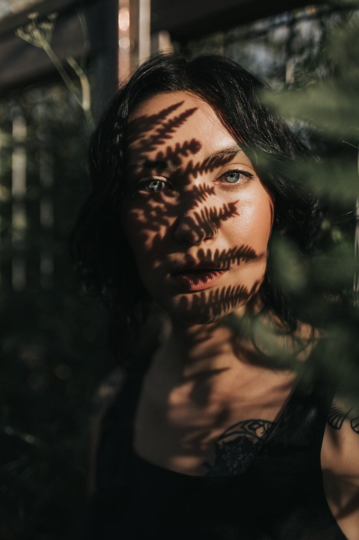 Woman with dark hair and light green eyes stares in to the camera surrounded by deep green ferns with a fern shadow over her face