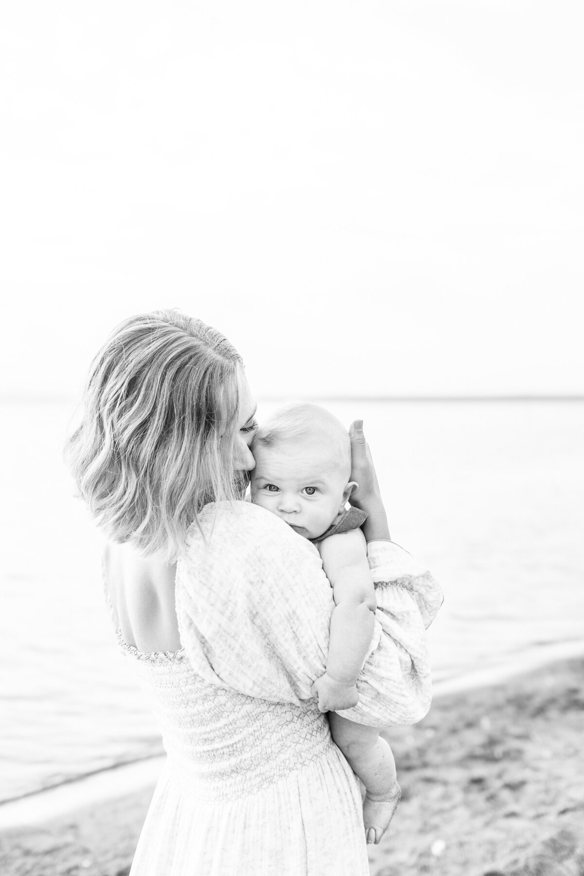 Black and white image of a mother kissing her baby while standing by the lake during their Kansas City milestone session.
