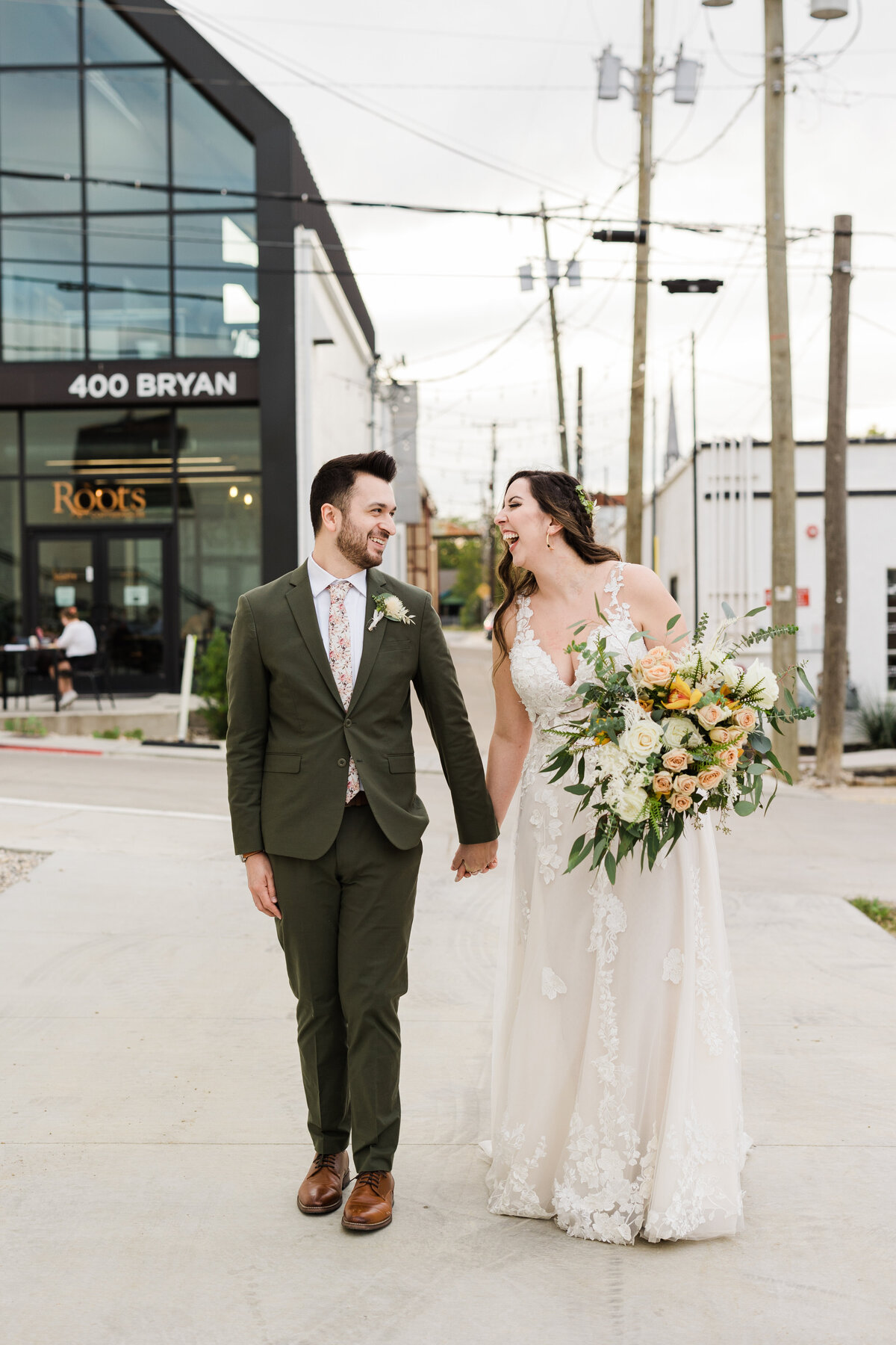 A portrait of a bride and groom walking outside and smiling at each other after their wedding at The 4 Eleven in Fort Worth, Texas. The bride is on the right and is wearing a sleeveless, intricate white dress with a large bouquet. The groom is on the left and is wearing a dark green suit with a floral tie and boutonniere.