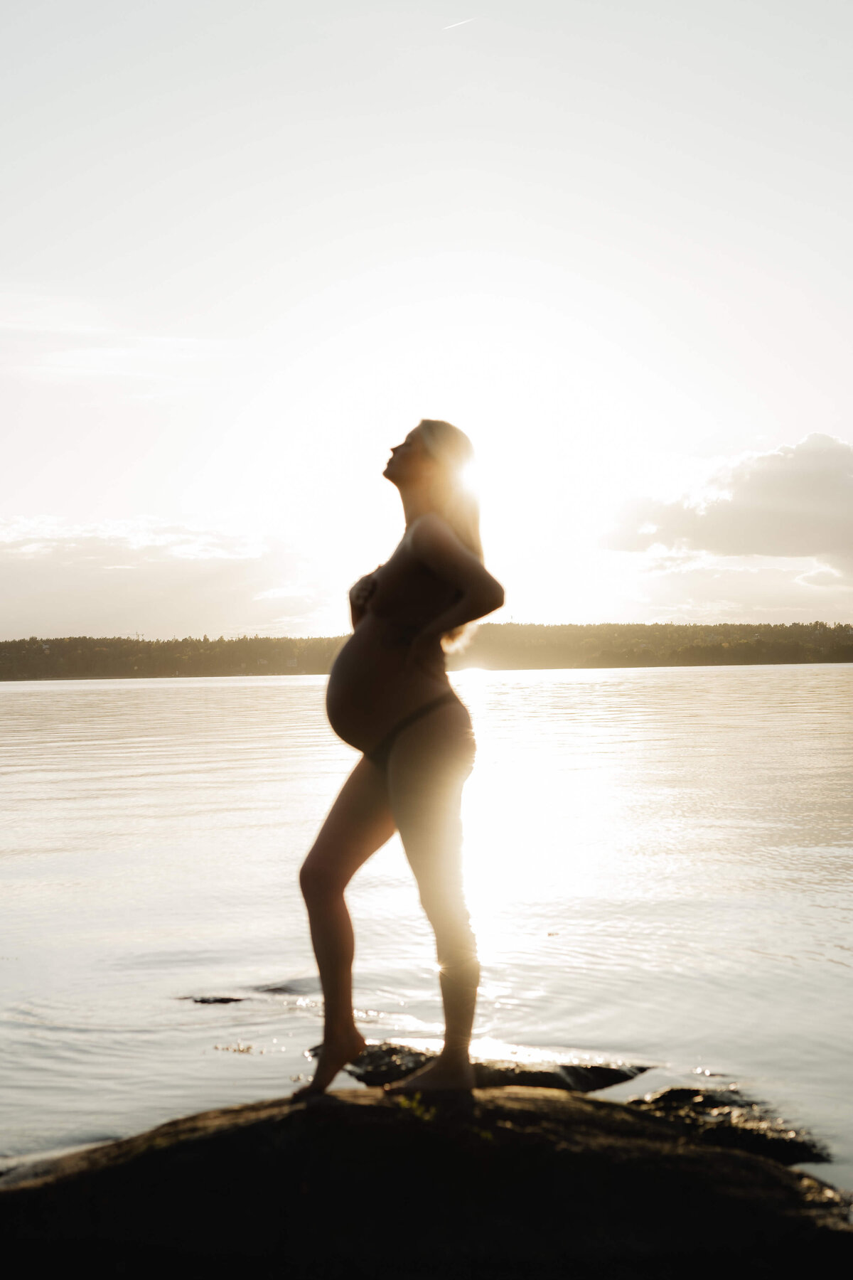 En gravid kvinna ståendes lutandes på sin ena höft ute vid vattnet under en gravidfotografering i Stockholm