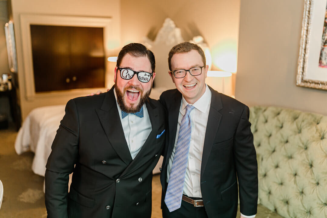 A groom with his arm around his groomsman standing and laughing in getting ready room