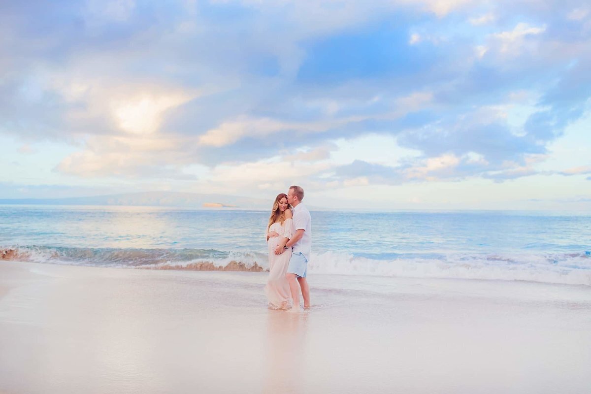Beautiful pastel tones surround couple embracing on the beach at sunrise for their family and maternity photography portraits at the beach in Wailea