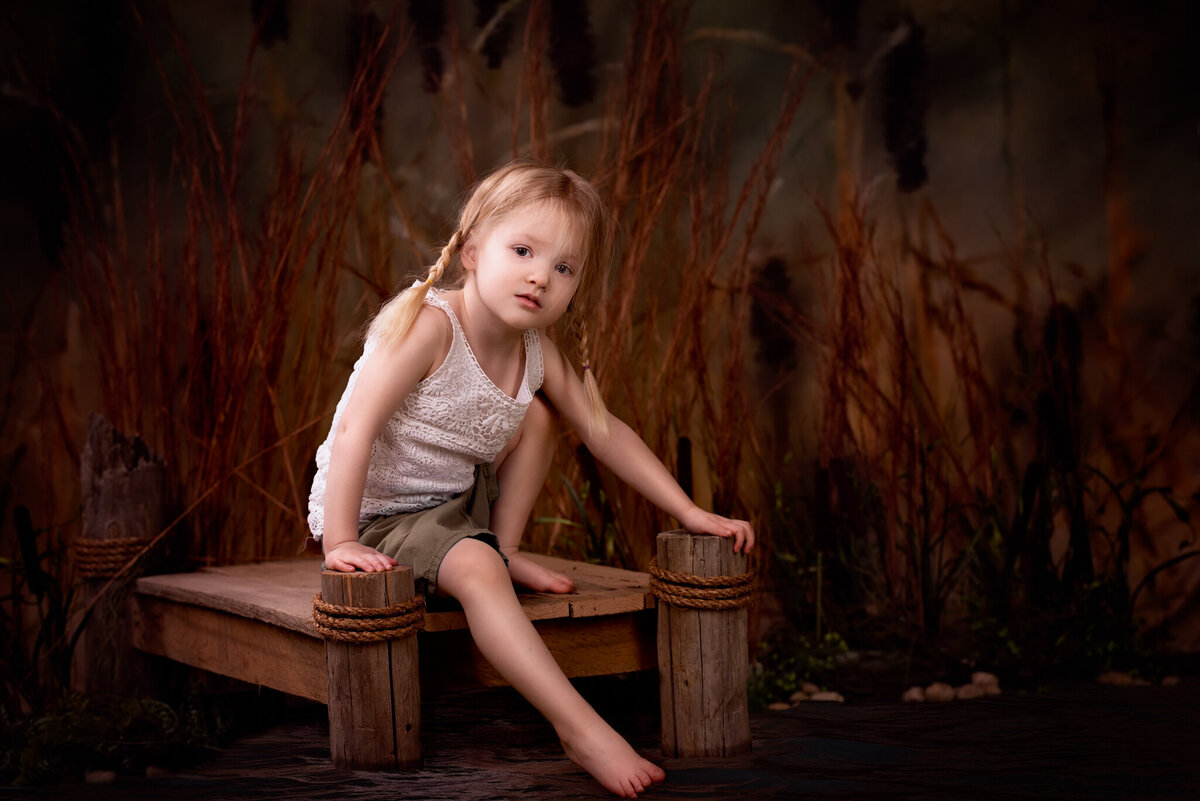 Fine art portrait of a young girl sitting on a wooden dock with a nature-inspired backdrop, captured in Overland Park, Kansas. A serene and natural option for families seeking rustic and artistic child photography.