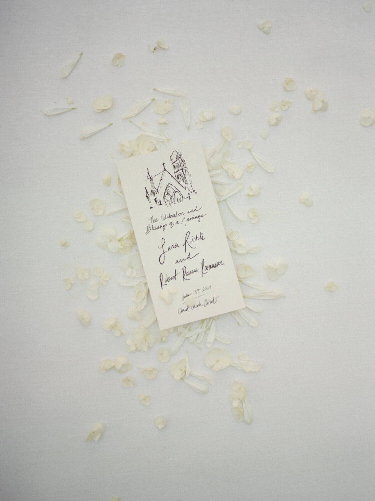 A soft, clean flat lay with white petals surrounding a timeless, gestural, hand-drawn wedding program for a unique Detroit wedding