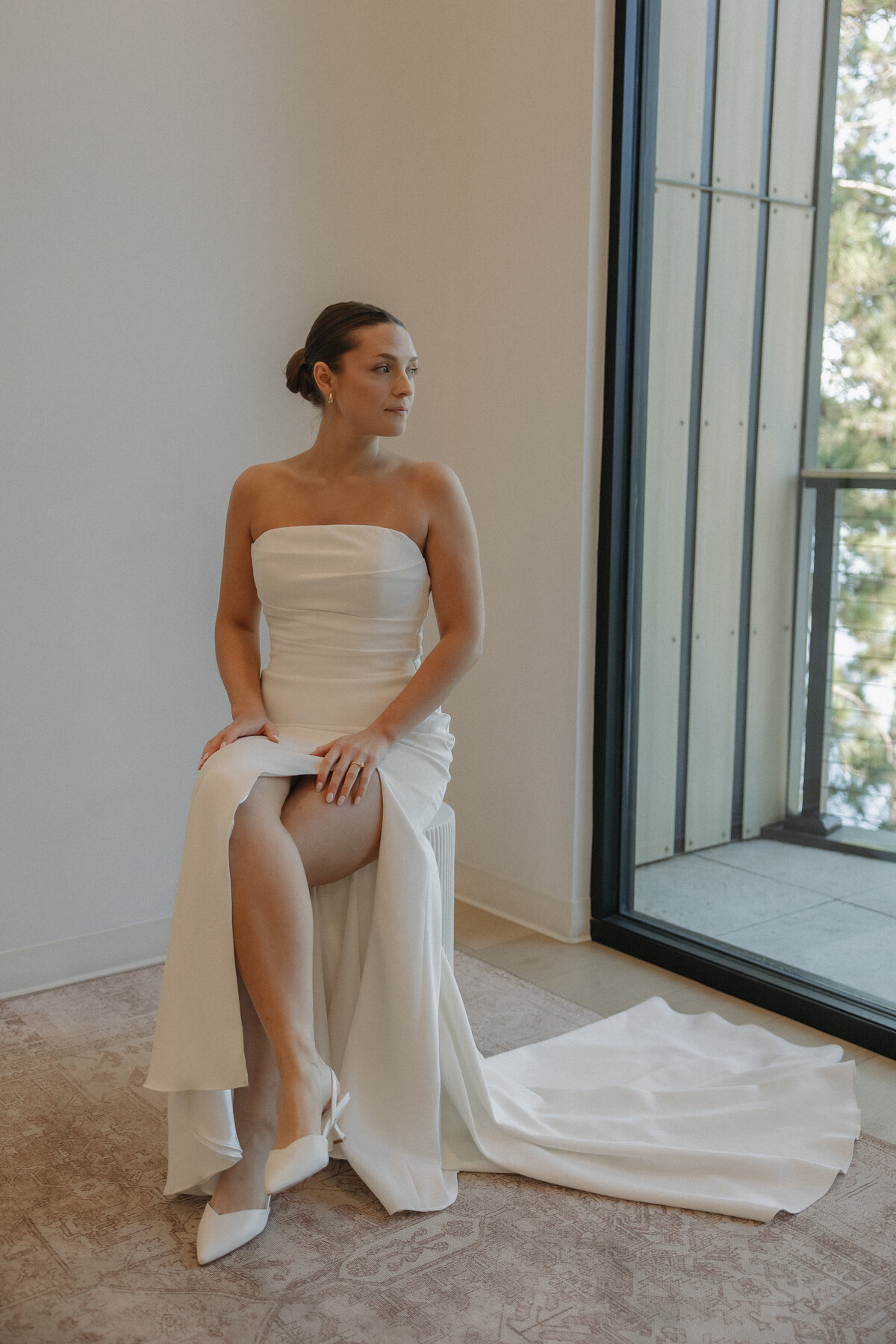 A bride in white satin strapless dress with a large leg slit sits on a cylindrical stool with both hands resting on her upper thigh. She looks over her left shoulder out a very large window that reveals a small balcony. Her face is stoic and she has a slicked back bun and small gold hoop earrings in.  