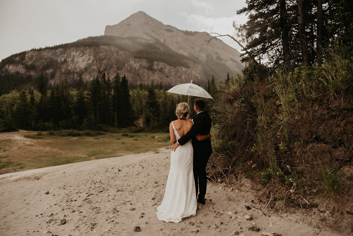 Bride and groom hugging under clear umbrella in the beautiful mountains, captured by Kelsey Vera Photography, intimate and romantic wedding photographer in Airdrie, Alberta. Featured on the Bronte Bride Vendor Guide.