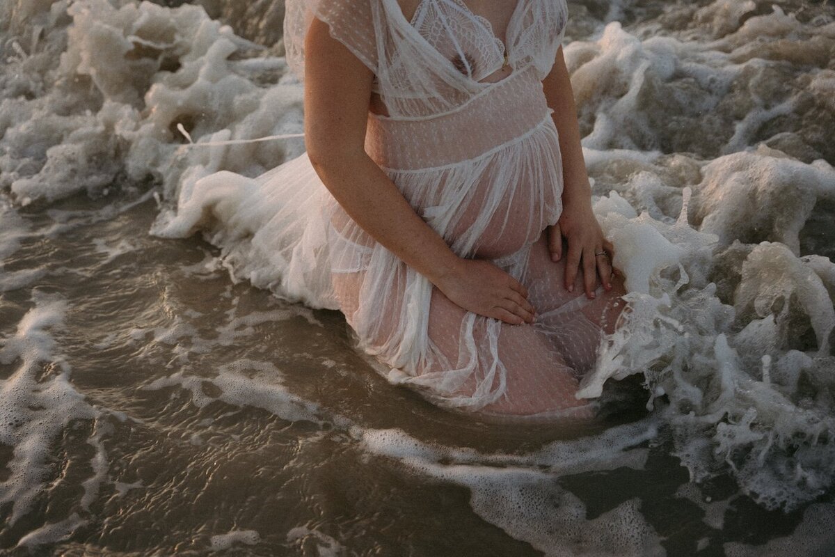Future maman à genou dans l'océan, robe légèrement transparente flottant au gré des vagues. Kamescop, séance grossesse artistique en Bretagne.