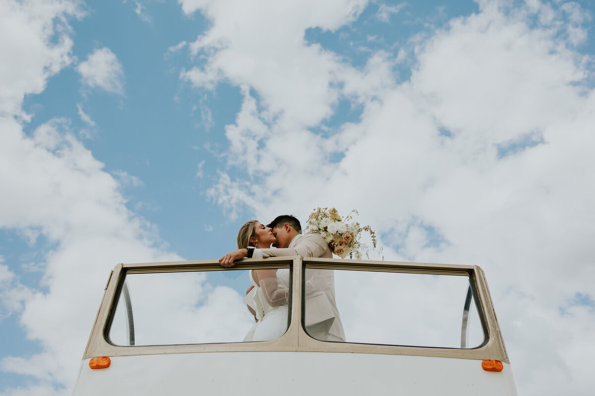 Groom kisses brides neck while they stand on top of double decker bus with blue clouded sky in background.