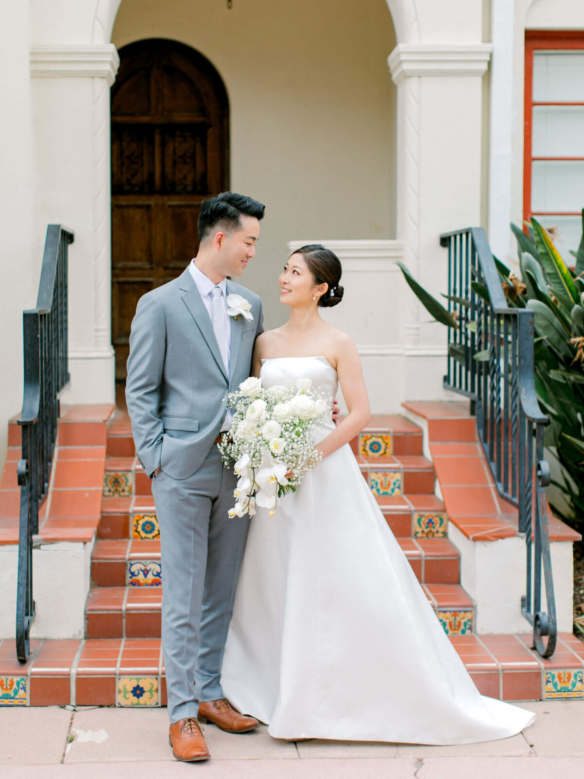 A smiling couple stands on tiled steps in front of an arched doorway, dressed in wedding attire. The bride holds a bouquet of white flowers.