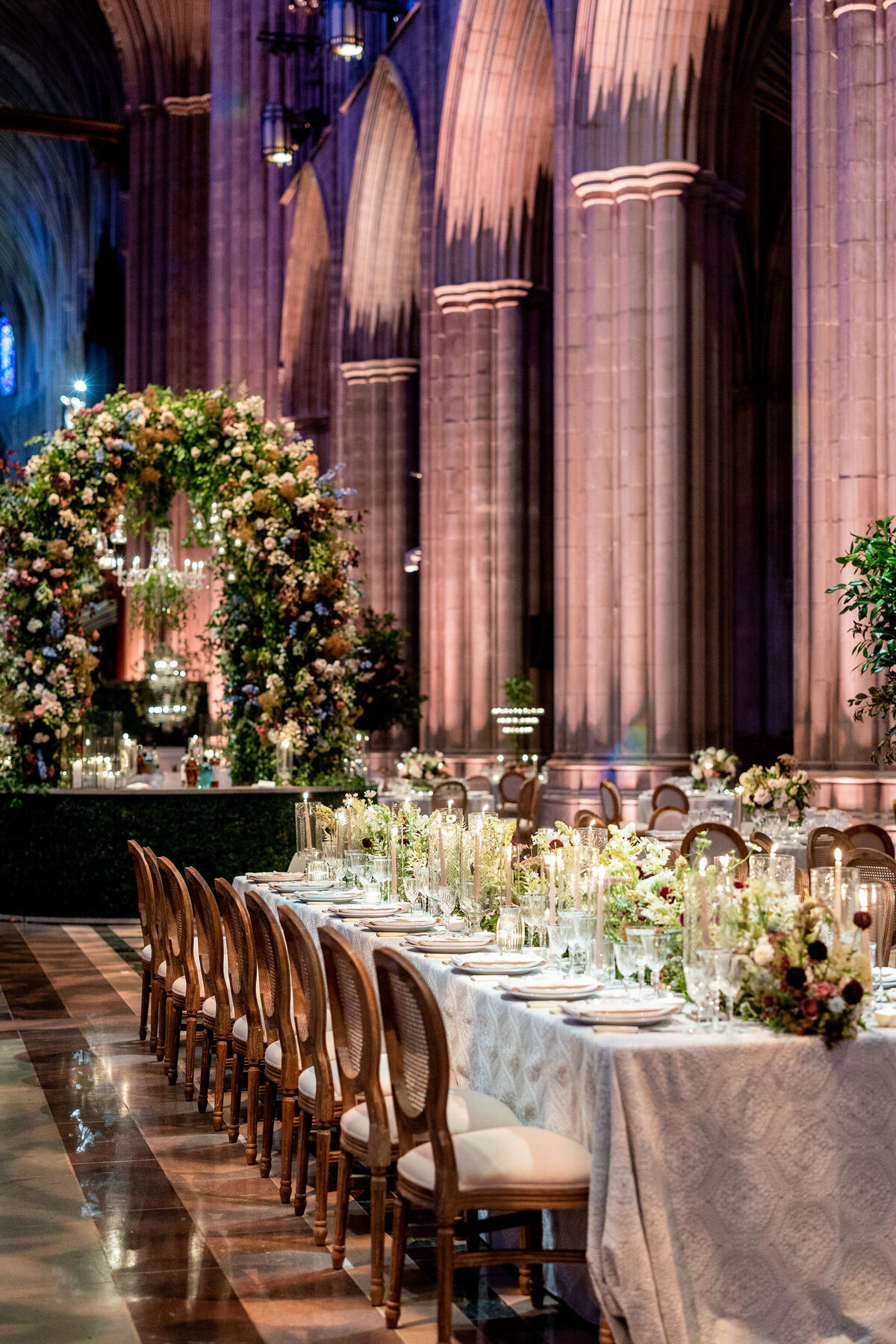 At the Washington National Cathedral in Washington D.C., photographer Twah Dougherty captured a beautiful moment where a bride and groom, both beaming with joy, walk hand in hand at their wedding reception. The bride, dressed in a stunning white off-the-shoulder gown, gracefully holds a bouquet of white flowers. Surrounding them, guests in formal attire cheer, clap, and snap photos to commemorate the happy occasion. The venue is adorned with elegant decorations, setting the perfect backdrop for the newlyweds' celebration. Additionally, Current Films provided videography services to capture the special moments of the day.