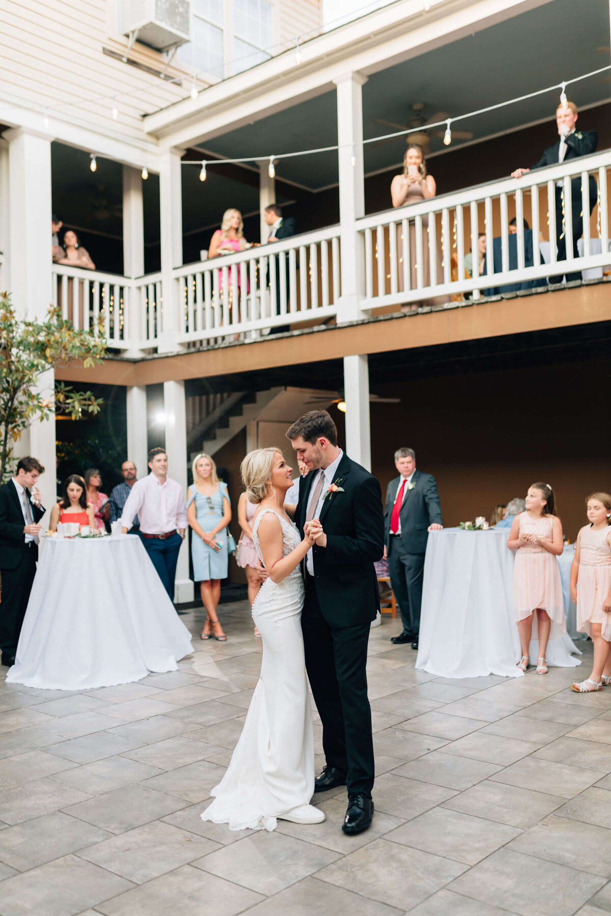 A bride and groom share their first dance at an outdoor wedding reception, surrounded by guests watching from both the ground and an upper balcony. The scene is festive and joyful.
