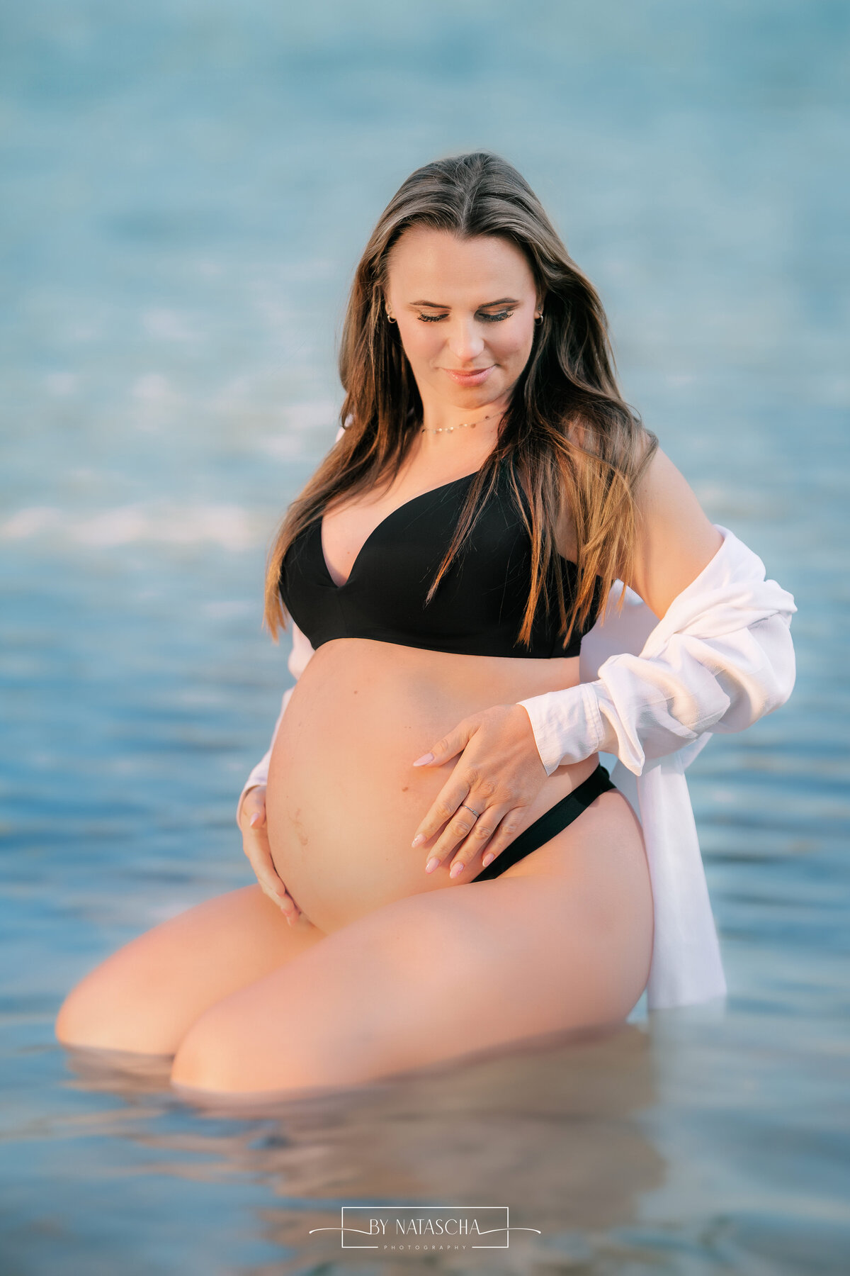 Zwangere vrouw in zwarte lingerie en blouse hangend over haar schouders, zittend op haar knie in het water en kijkend omlaag in Berghem, Herperduin in het blauwe water