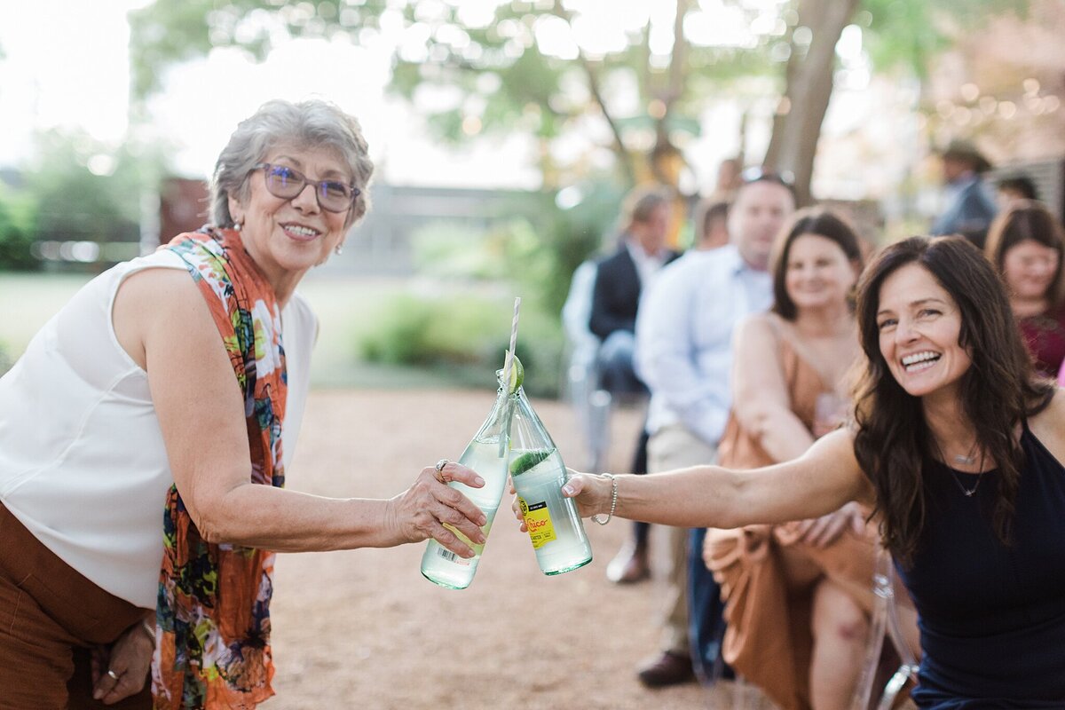 Two wedding guests toast with bottles of Topo Chico across the ceremony aisle right before an outdoor wedding ceremony at Artspace111 in Fort Worth, Texas. Both guests are smiling and looking at the camera as the bottles clink and many guests can be seen behind them in the background.