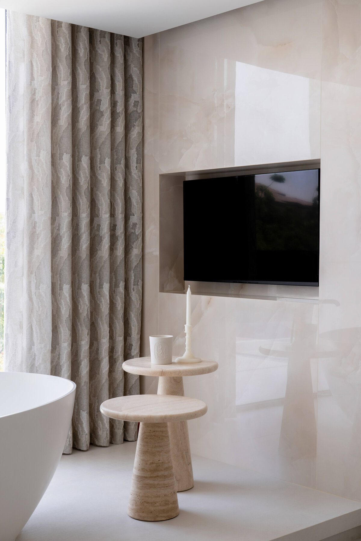 Television recessed into the light marble wall next to the tub in a chic primary bathroom.