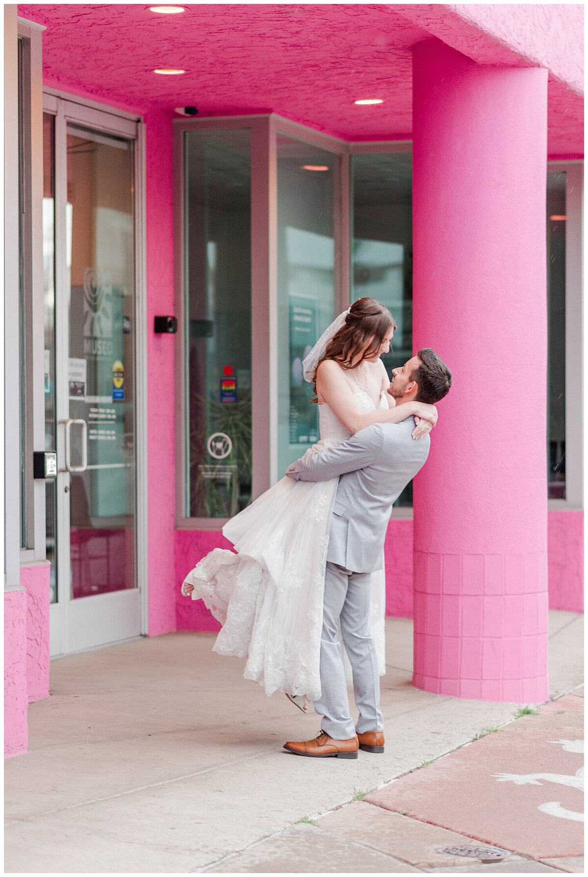 A bride picks up his new bride, both of them in wedding attire while he spins her and they look at each other in front of a bright pink building with a large pillar outside of skylight venue