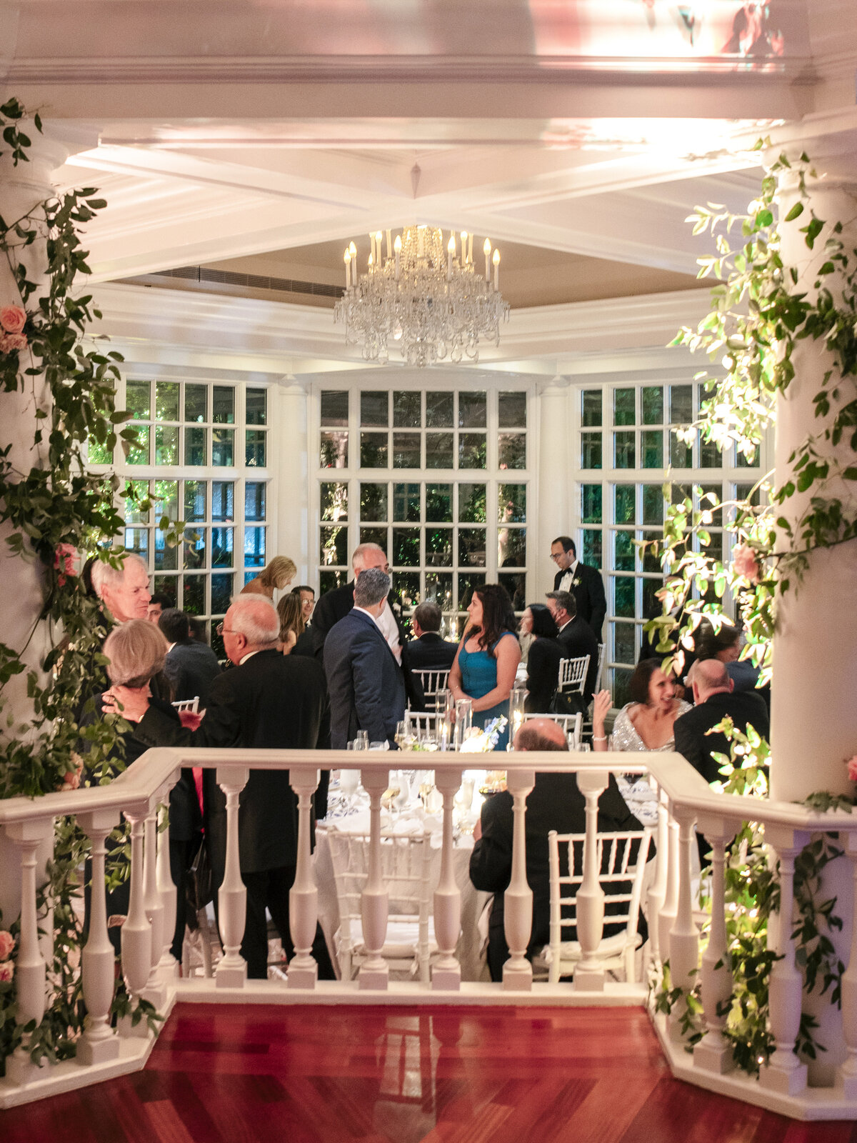 A formal gathering in a bright, elegant room with a large chandelier and floor-to-ceiling windows. Guests in evening attire are seated at tables adorned with white linens. The room is decorated with greenery and soft lighting for a festive atmosphere.