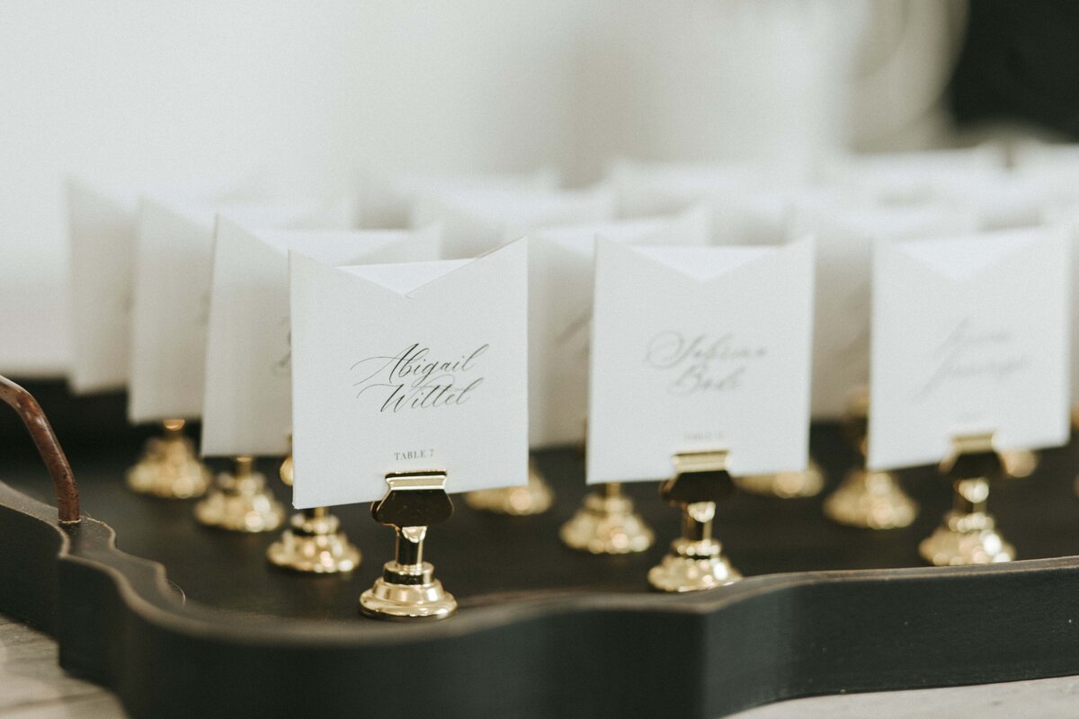 White wedding place cards with black cursive font on gold place card holders atop a dark green tray.