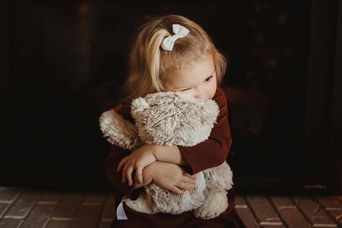 A little girl is hugging her teddy bear while sitting in front of the fireplace.