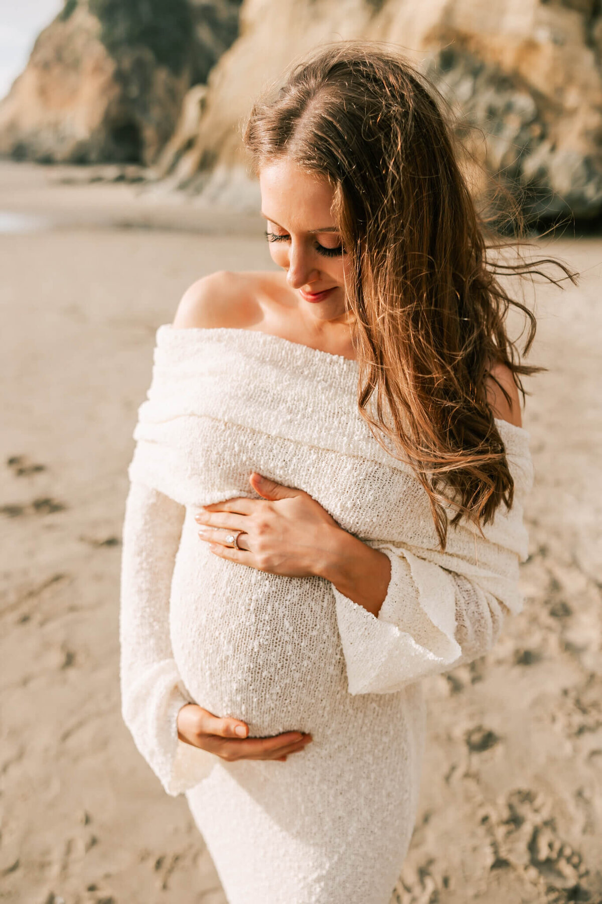 portrait of a woman on the beach with her hands on her belly. She has long brown hair and is smiling down at her belly. She is wearing a cream dress  that is off the shoulder.