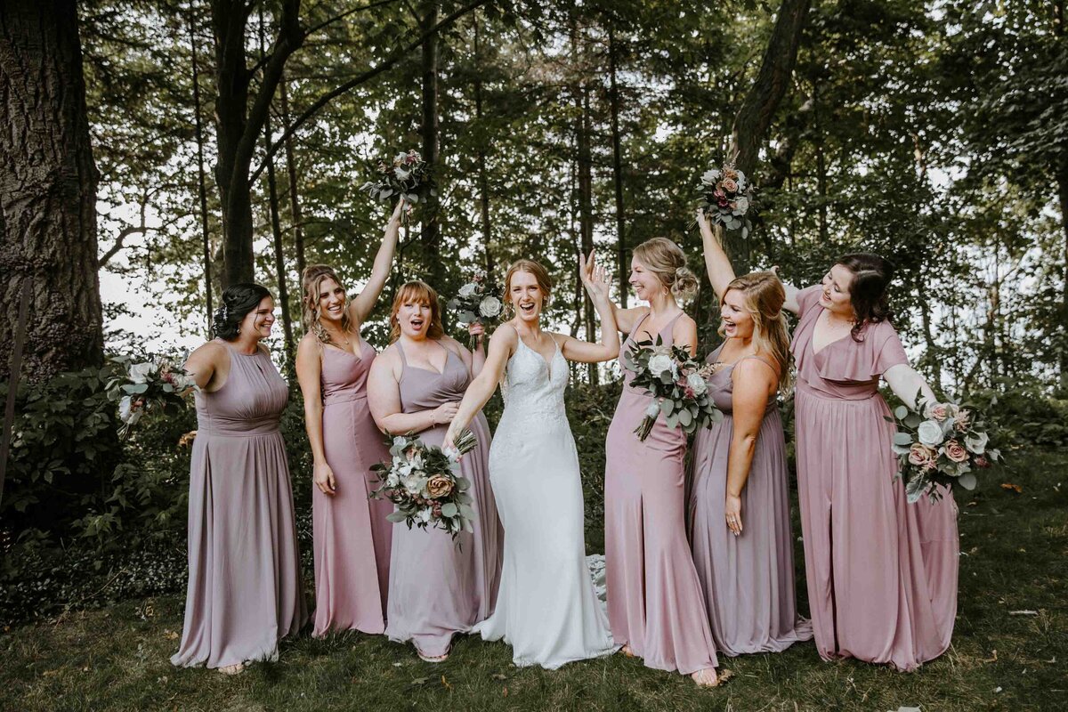 Bride and her bridesmaids pose for wedding day photos against treeline in Exeter, Ontario. Bridesmaids in long gowns with various pink and purple hues. All are throwing their hands up in celebration.