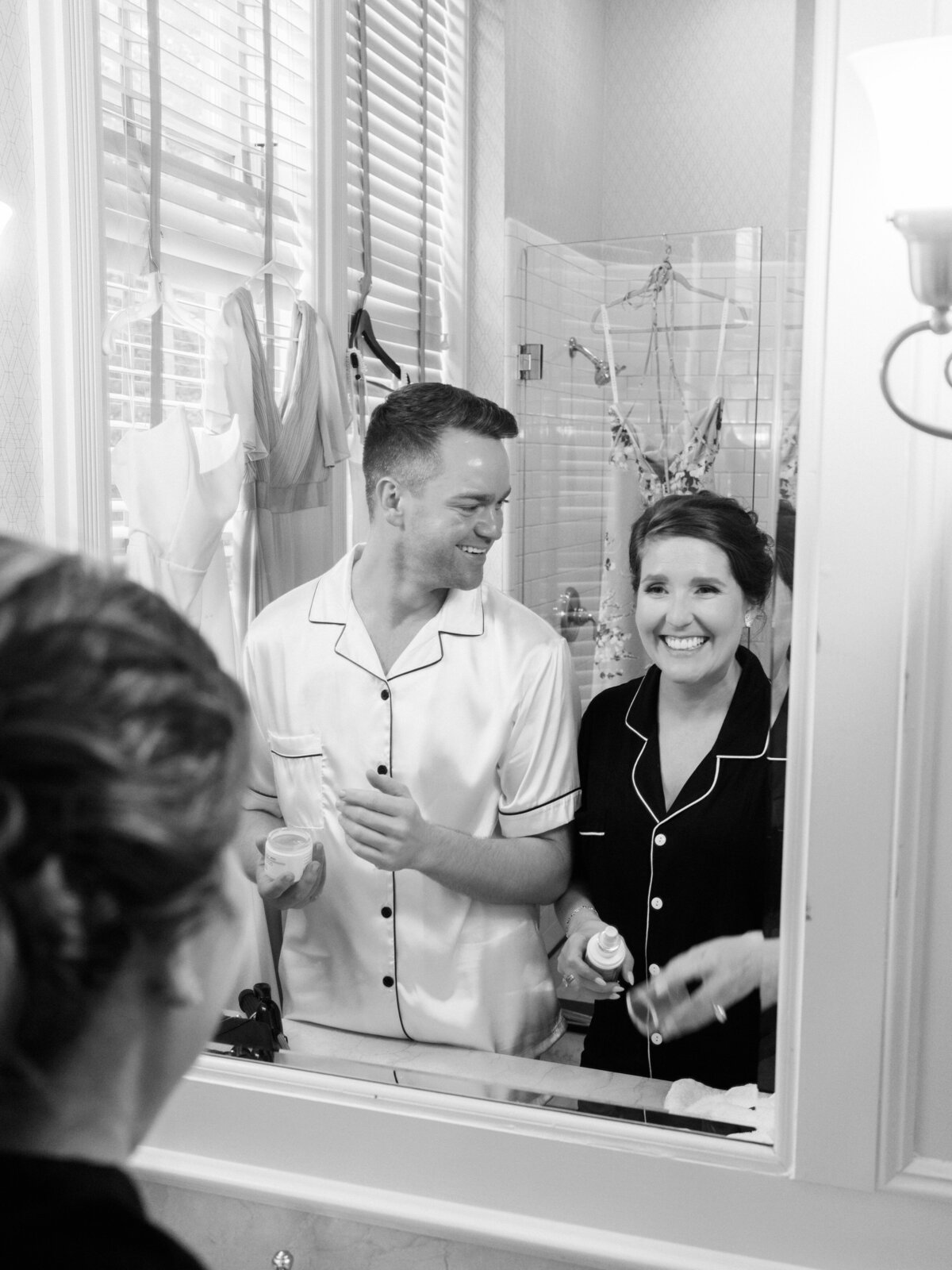 A joyful moment captured as the groom and a bridesmaid smile at each other in the mirror, reflecting excitement and intimacy in the context of wedding preparations.