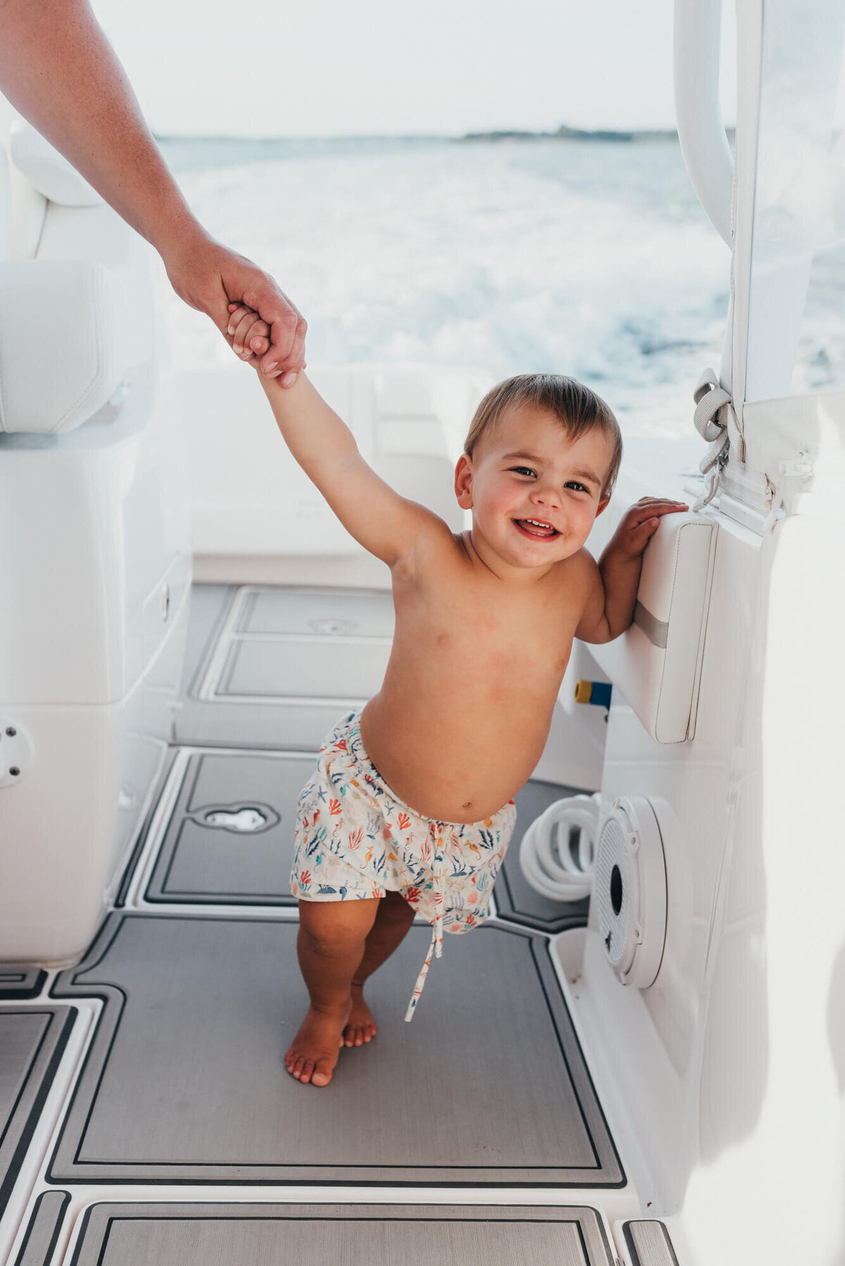 A toddler boy wearing an ocean themed bathing suit holds on to his father's hand while his father drives their boat during their photoshoot with Boston Family Photographer, Allison Wolf