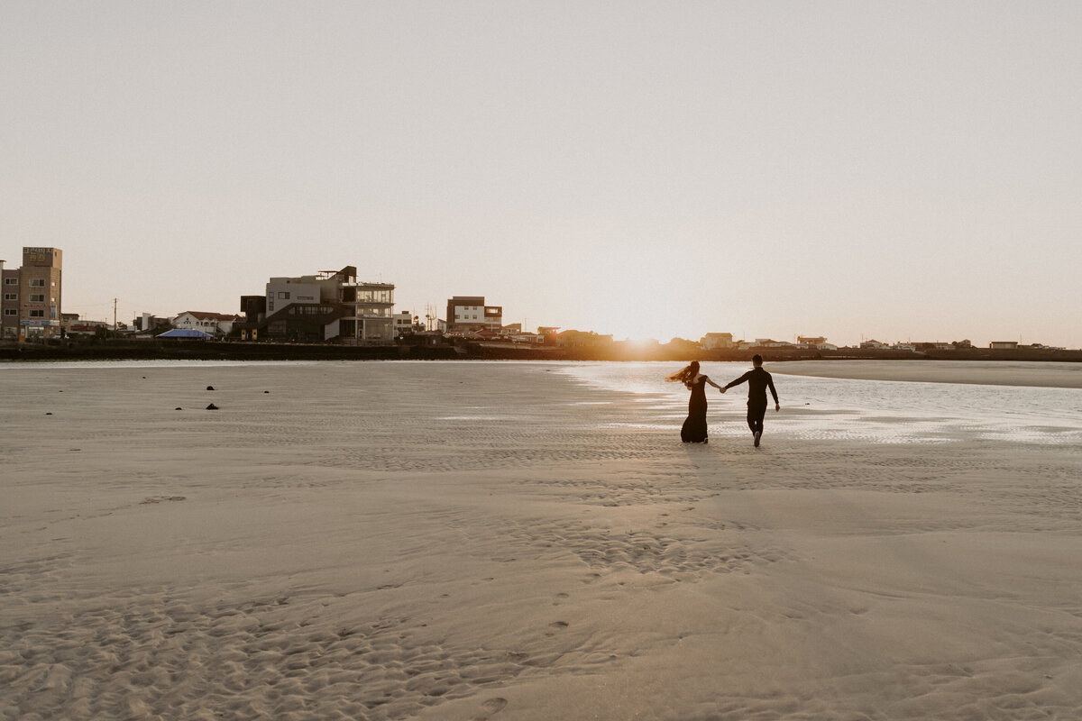 the couple holding hands while running in the shore under the golden sunset of Jeju Island
