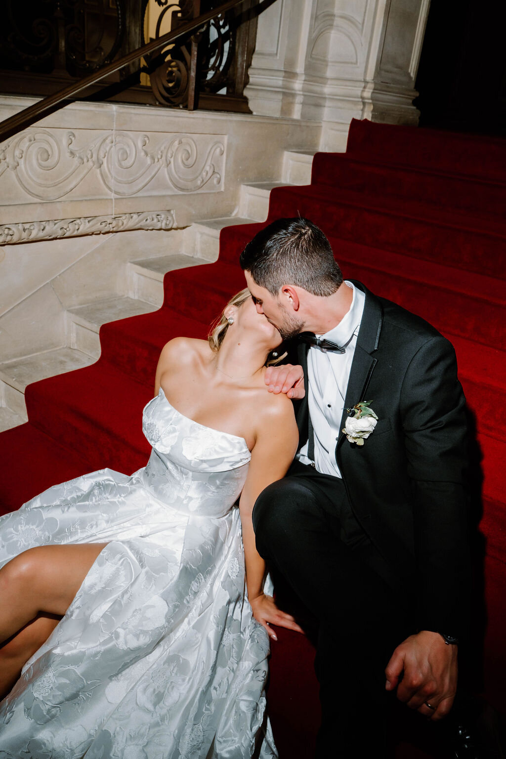 Rhode Island Wedding Photographers | A couple dressed in formal attire shares a kiss while seated on elegant red carpeted stairs. The setting is romantic and intimate.