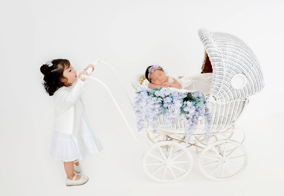 Toddler gently holding a stroller while a sleeping baby rests inside with chin on arms, surrounded by cascading lilac flowers.