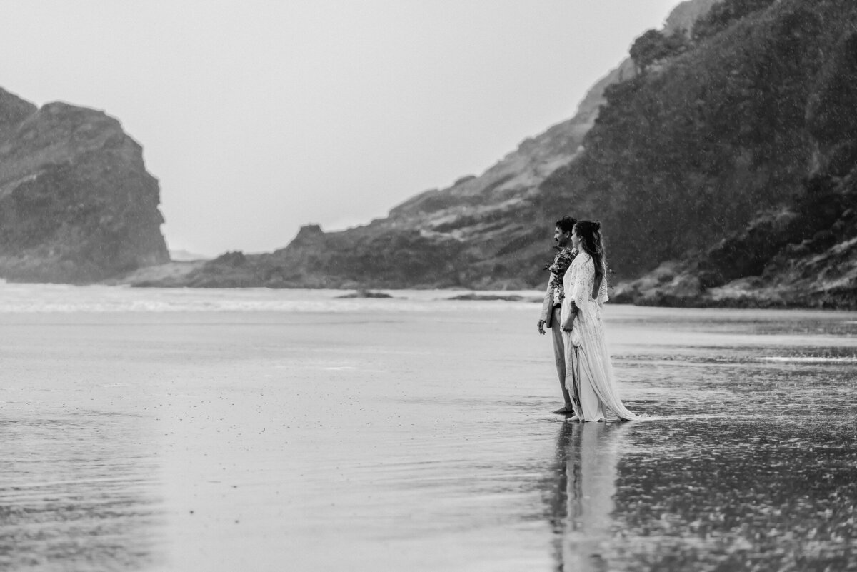 During their Oregon coast elopement, a couple gazes out towards the ocean. The rain is pouring on them and the waves are rolling in.