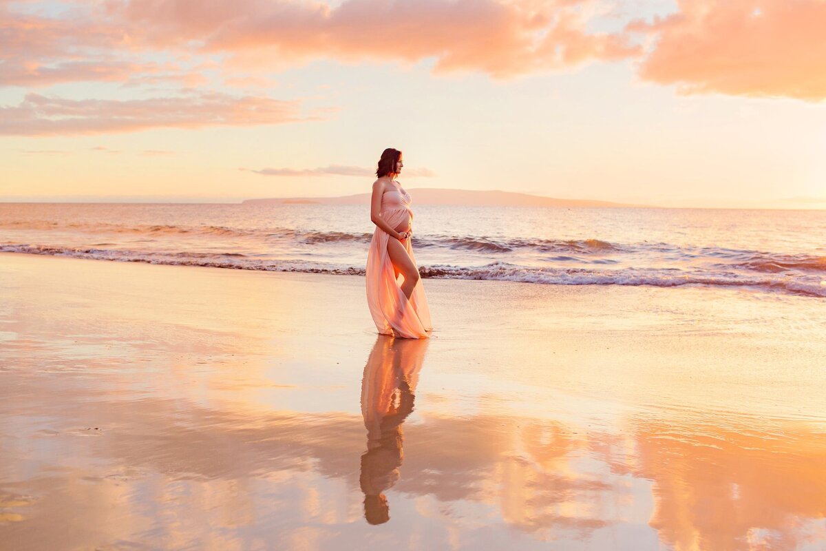 Pregnant beautiful redhead in pink gown holds her belly while reflective sand enhances her babymoon portraits by Love + Water