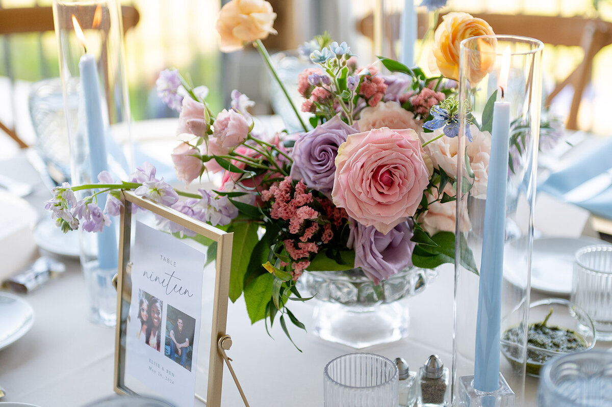 Elegant reception centerpiece in a glass compote vase featuring pastel flowers including pink and lilac roses, greenery, and light blue taper candles on a white table setting.