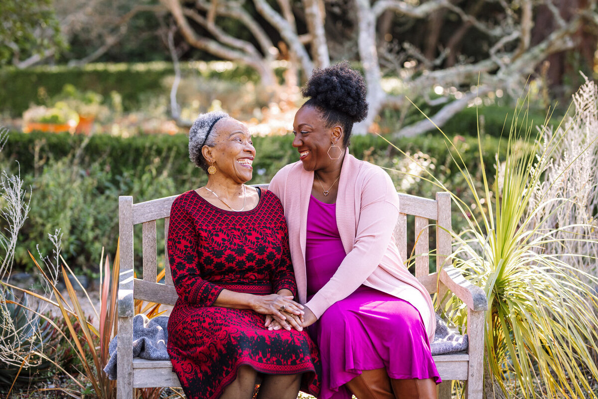pregnant woman sitting on a bench with her mother with both holding hands and looking at each other smiling at a Berkeley garden