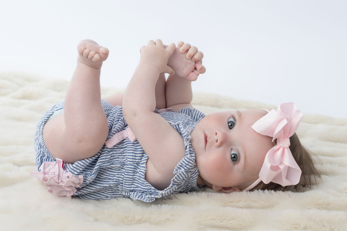 A baby lying on back on a cream blanket, wearing a striped outfit and pink bow, holding feet.