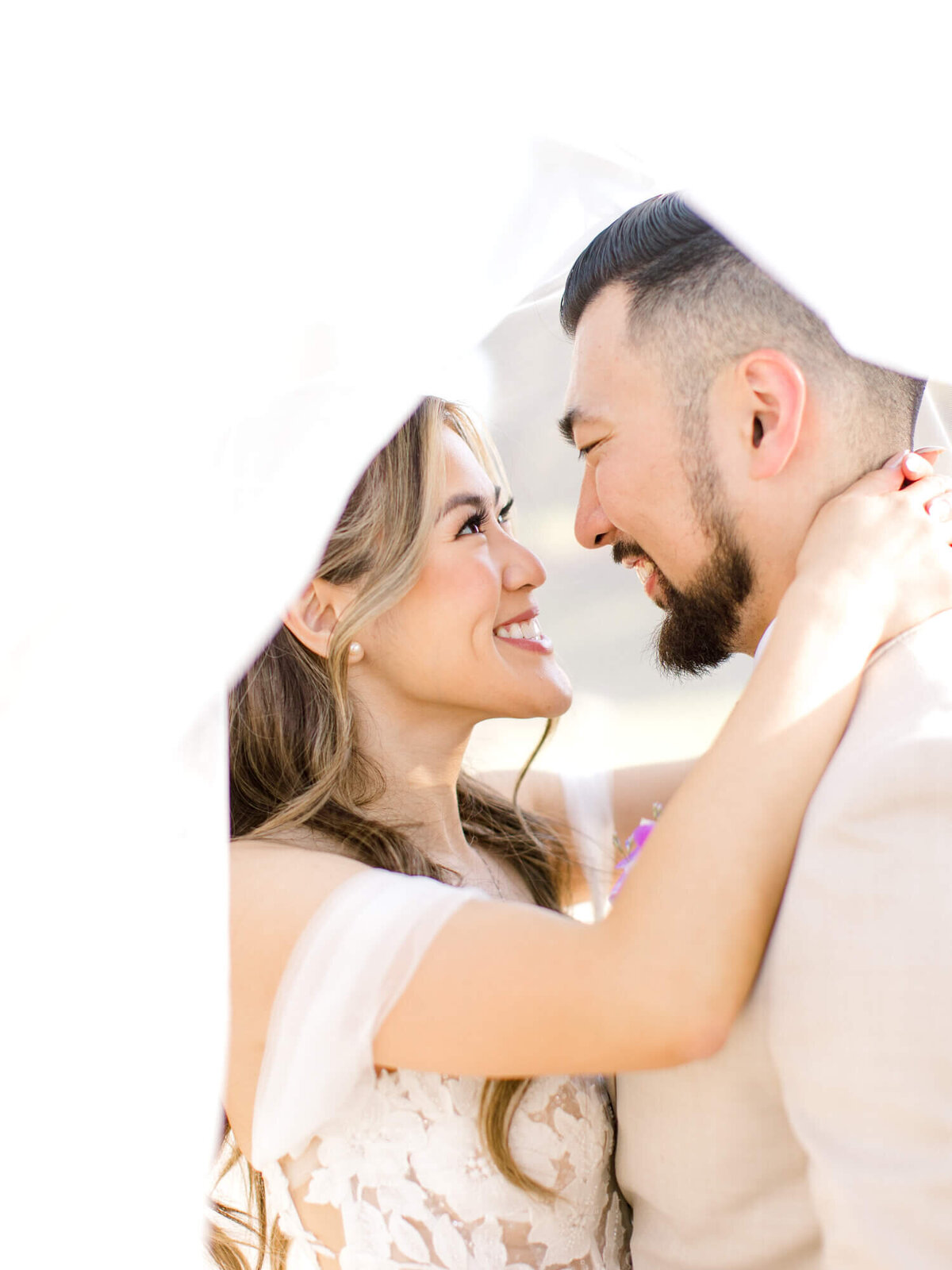 A bride and groom share an intimate moment under a flowing veil, gazing into each other's eyes and smiling. The atmosphere is romantic and joyful.