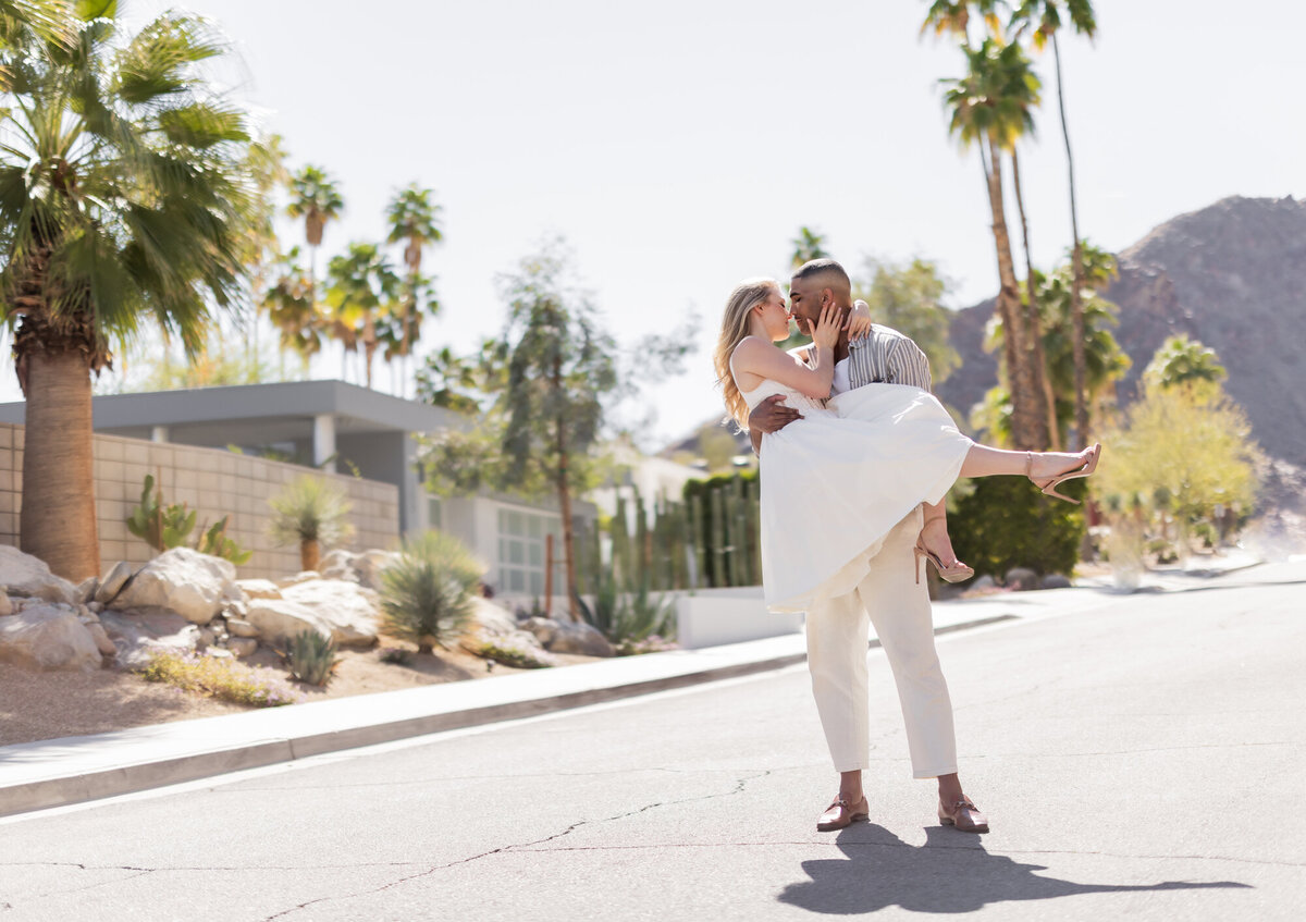 A romantic and cinematic moment in Palm Springs, California, as a couple shares a kiss with the desert landscape and iconic mid-century modern architecture in the background. The groom-to-be lifts his partner in an intimate embrace, creating a timeless engagement or anniversary portrait.
