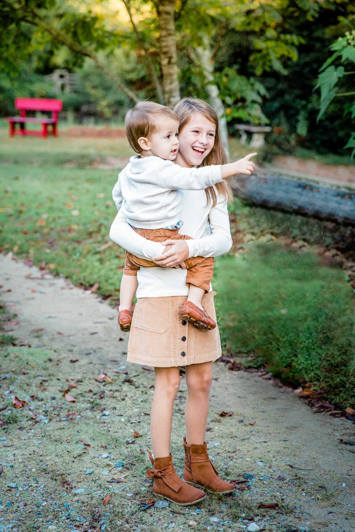 A sister smiles as she holds her baby brother while he points to something in the distance.