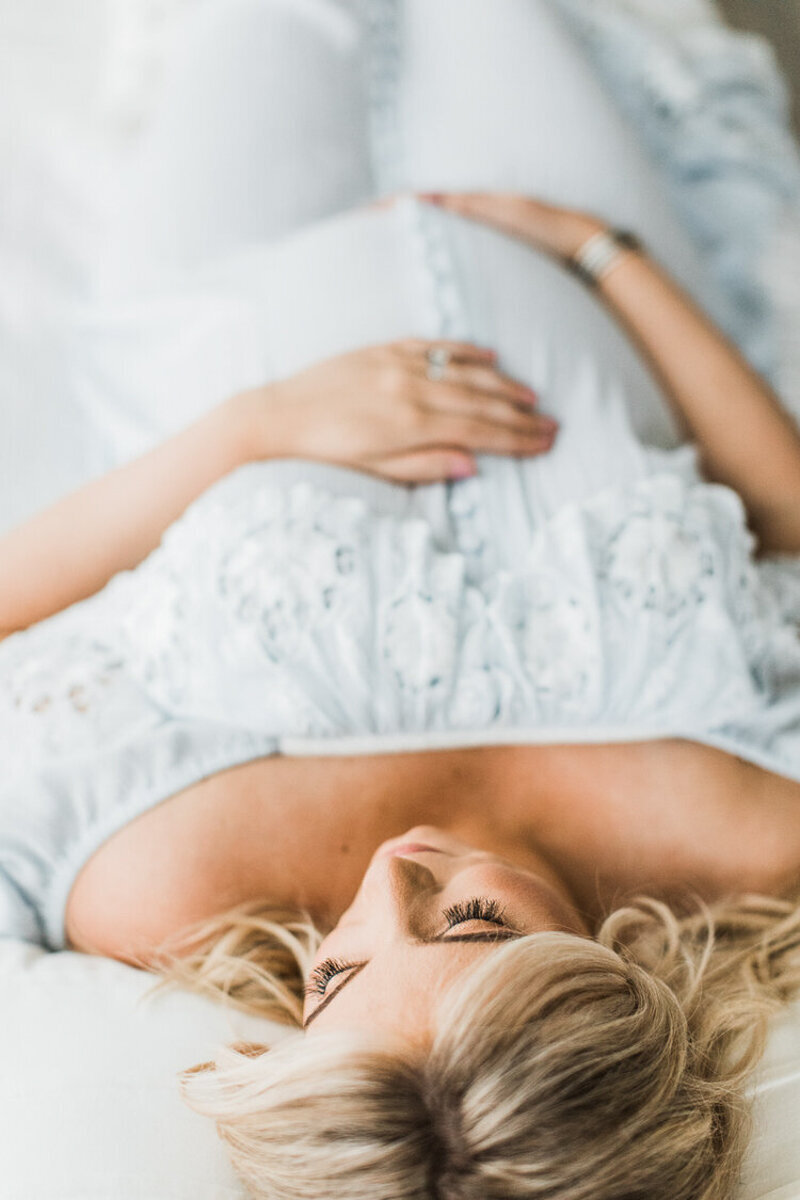 pregnant mom rests by laying down on the bed with blond hair long lashes and blue dress