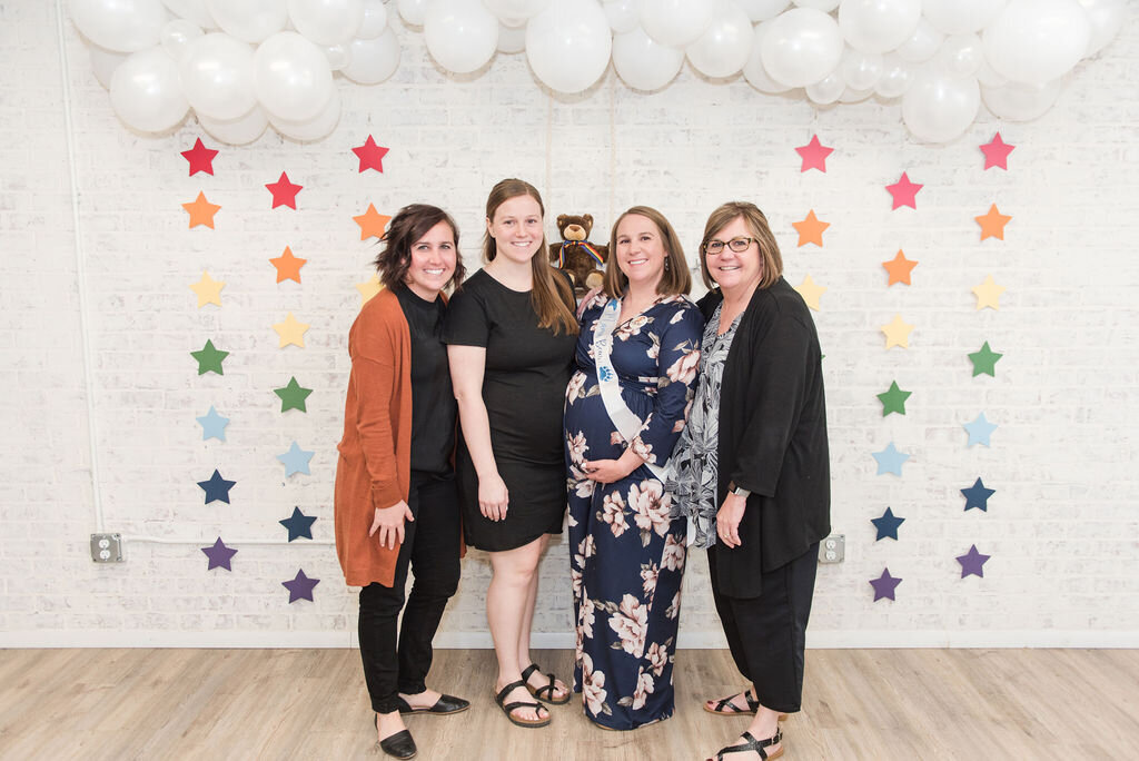 Mom-to-be standing with her sisters and mother against a rainbow star garland and white balloon cloud backdrop at a baby boy woodland shower.