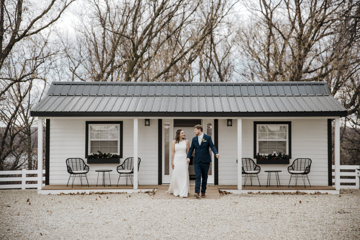 A newlywed couple holding hands in front of a charming white cottage with black accents, celebrating a moment of togetherness in a quaint and peaceful setting taken by jen Jarmuzek photography a Minneapolis wedding photographer