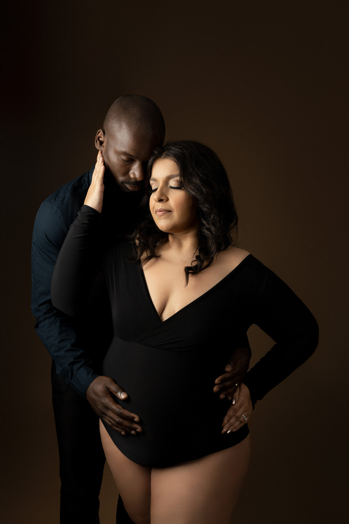 Expectant mom and dad in London, Ontario studio maternity session mom and dad are slightly angled to the camera with dad behind mom. Mom and Dad's heads are gently touching and Dad is holding mom's baby bump.