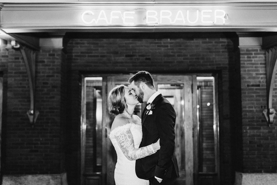 A bride and groom pose under the Cafe Brauer sign for a wedding portrait at night