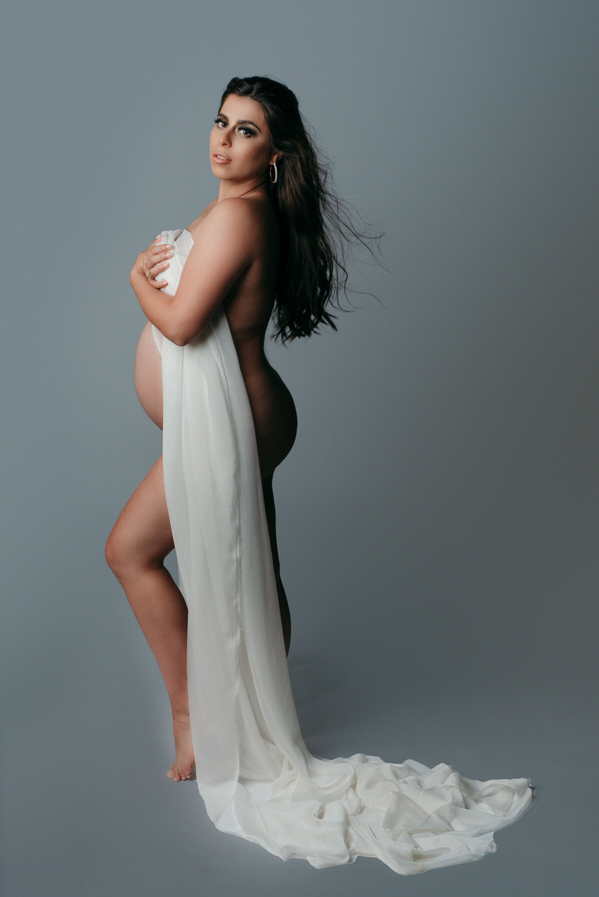 Woman standing in maternity pose holding white fabric draped over baby bump