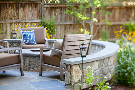 Cozy outdoor seating area with a curved stone fire pit, wooden chairs, and a decorative pillow, surrounded by lush greenery and a wooden fence.