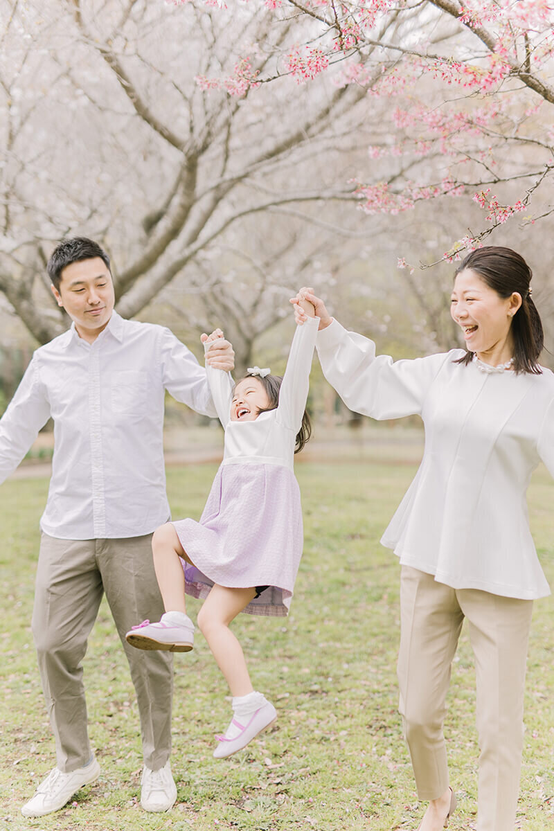 ママとパパと手をつないで、ジャンプしながらニッコリ笑っている女の子の可愛い写真。桜がバックに映えている、オシャレな家族写真。相模原市でYukika Ishikawa Photographyが撮影。
