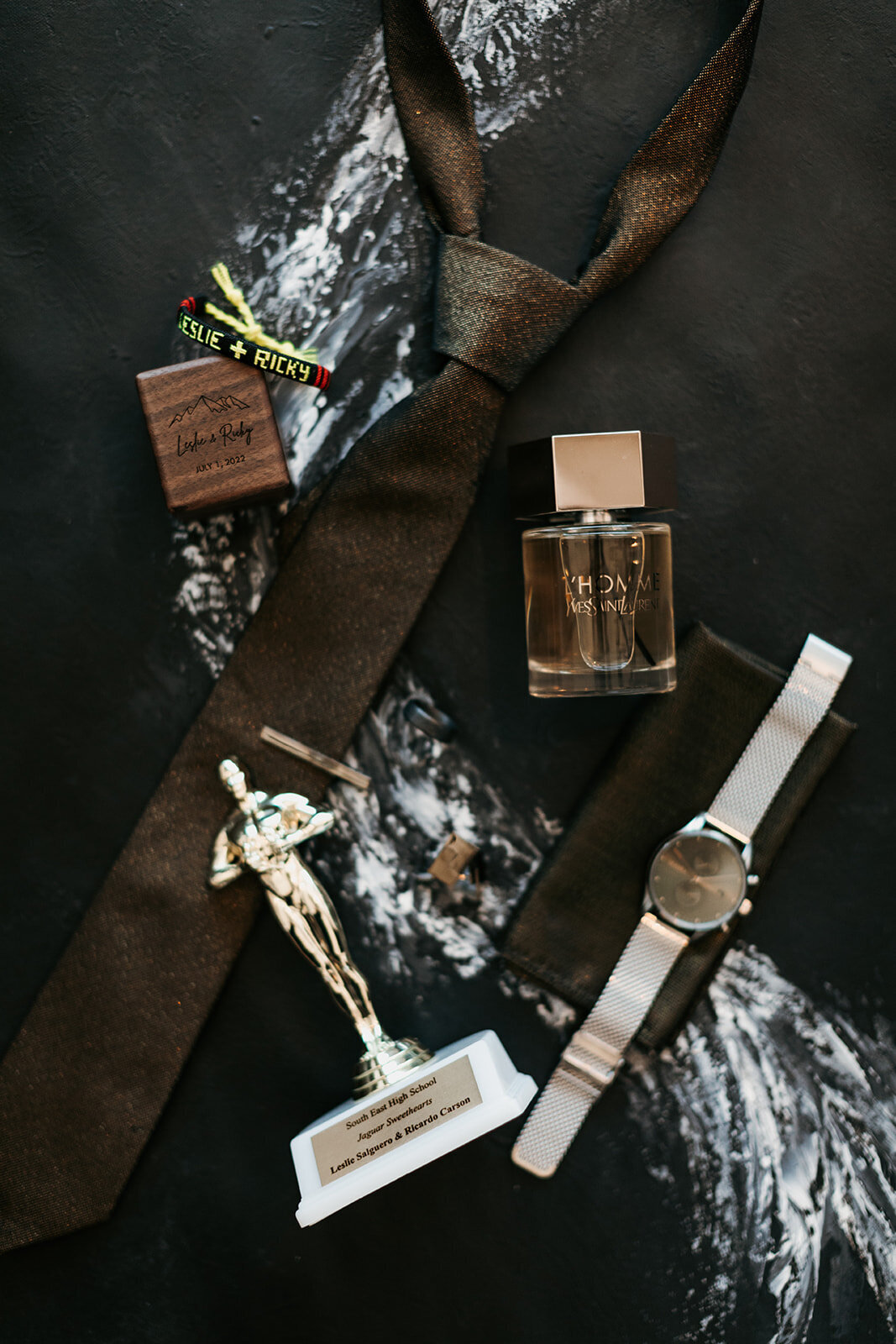 A flat lay of groom's accessories, including a dark tie, wristwatch, Yves Saint Laurent cologne, custom bracelet with "Leslie + Ricky," and a small trophy labeled "South East High School.