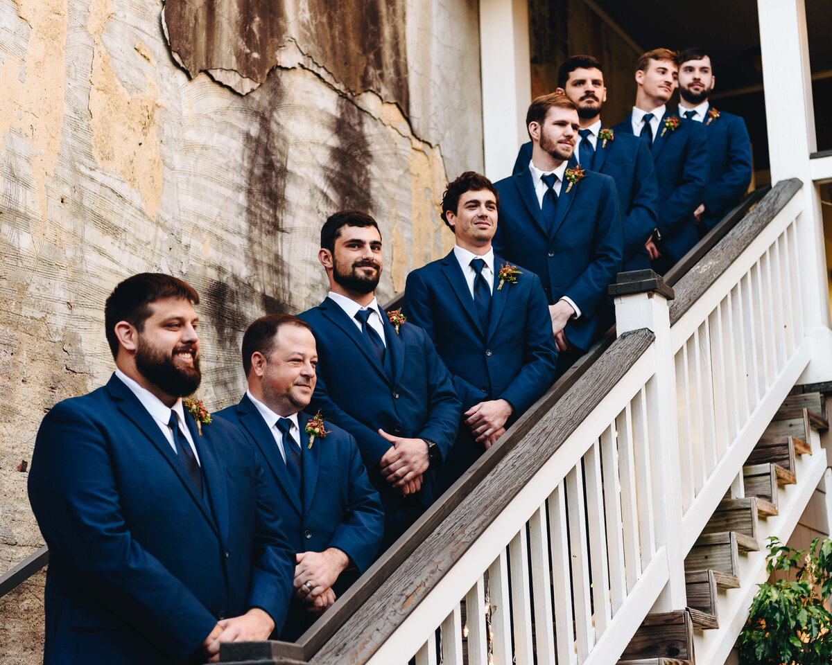 Eight groomsmen in blue suits stand in a line on an outdoor staircase beside a weathered, textured wall, smiling and looking ahead.