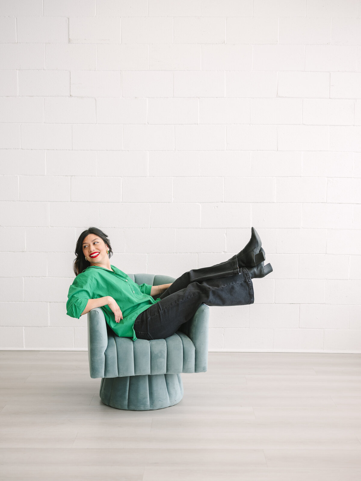 Woman laughing in green blazer and boots while reclining in teal chair during NYC brand session.