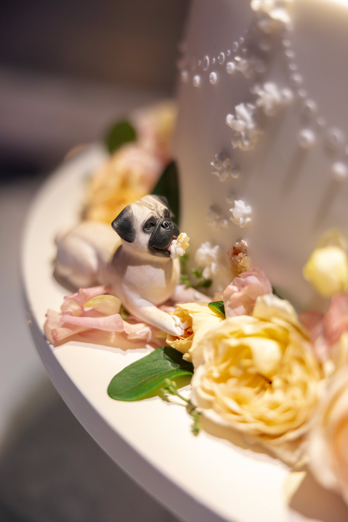 A detailed cake decoration featuring a small pug figurine surrounded by yellow and pink flowers. The pug, made from edible material, is resting on the cakes surface, adding a playful touch to the elegant floral arrangement.