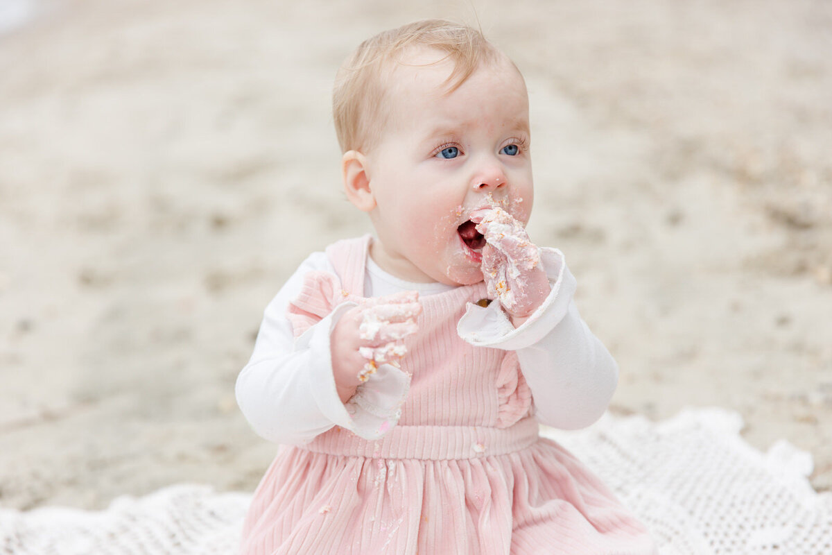 One-year-old girl smiles while licking icing off her hands on the beach during milestone session
